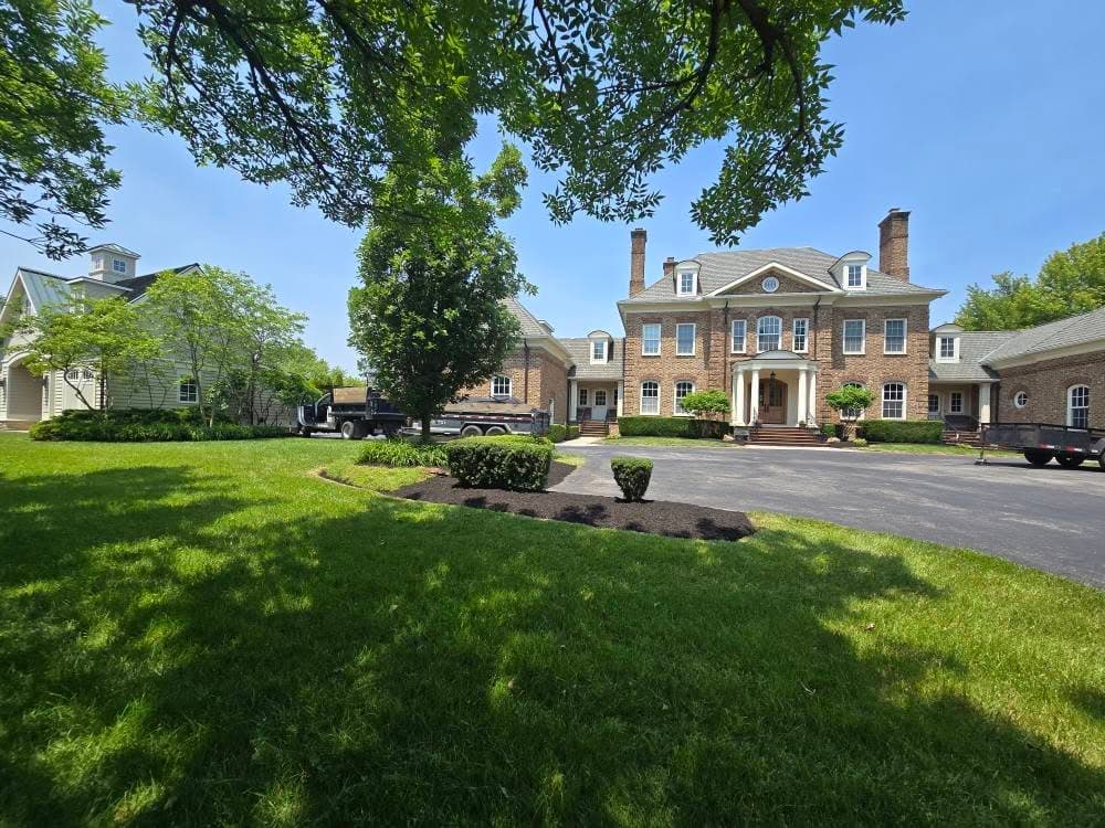Elegant brick mansion with green lawns, trees, and a paved driveway on a sunny day.