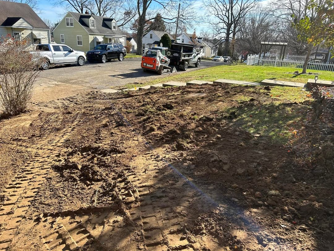 Excavated yard with tire tracks, construction equipment, and residential houses in background.