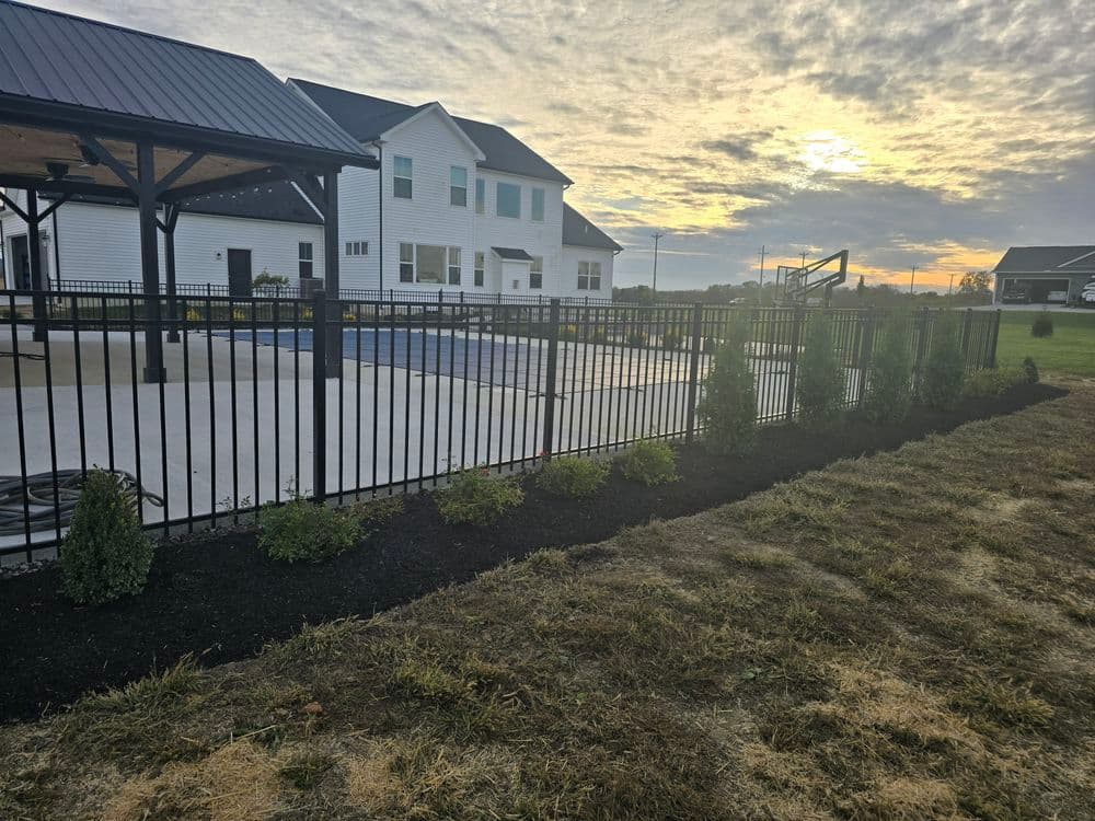 Black metal fence surrounding a home with a pool at sunset, landscaped with shrubs.