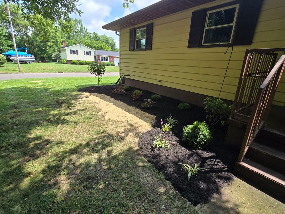 Landscaped yard with freshly mulched flower beds beside a yellow house and green grass.