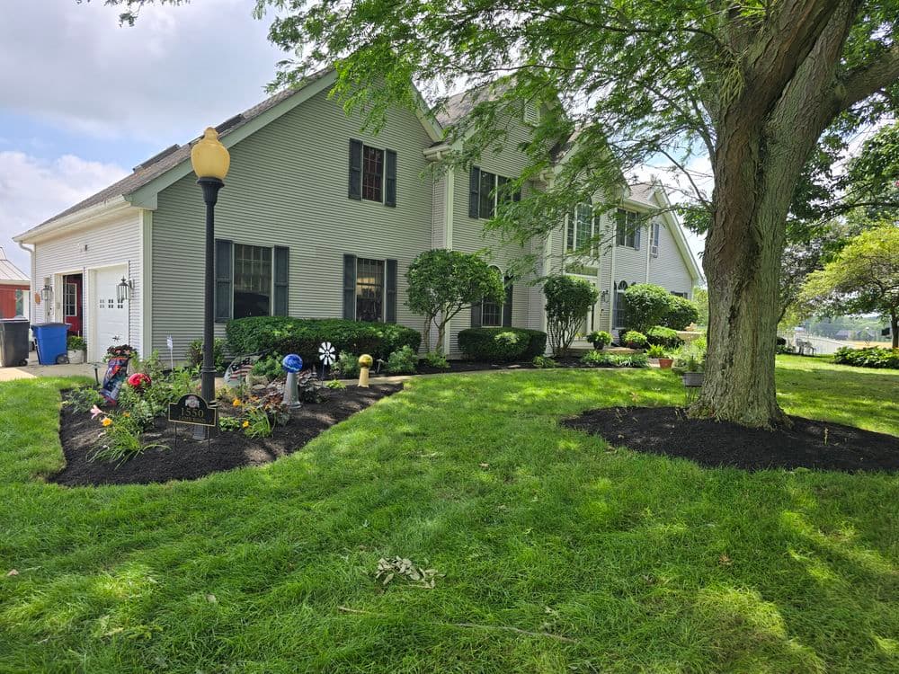 Charming two-story home with landscaped yard and large tree, featuring flower beds and garden decor.