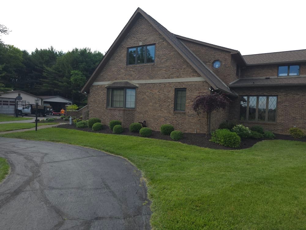Brick house with landscaped yard and driveway, surrounded by trees and greenery.