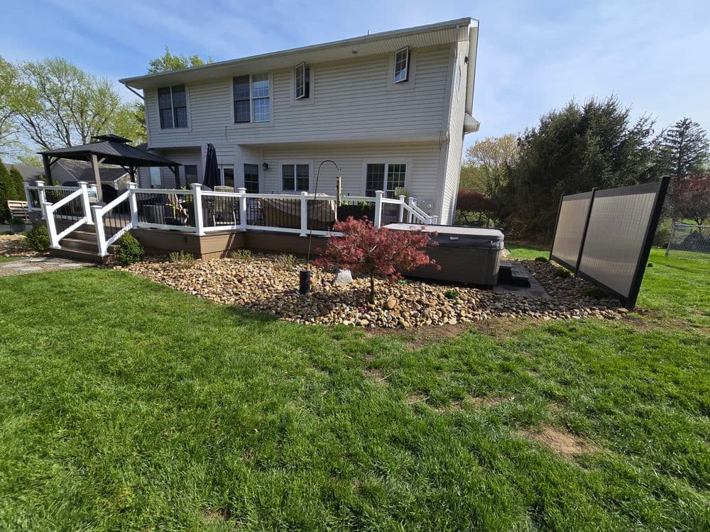 Backyard patio with hot tub, decorative rocks, and landscaping beside a two-story home.