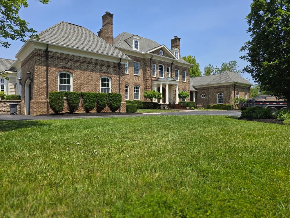 Elegant brick mansion with lush green lawn and well-trimmed hedges under a clear blue sky.