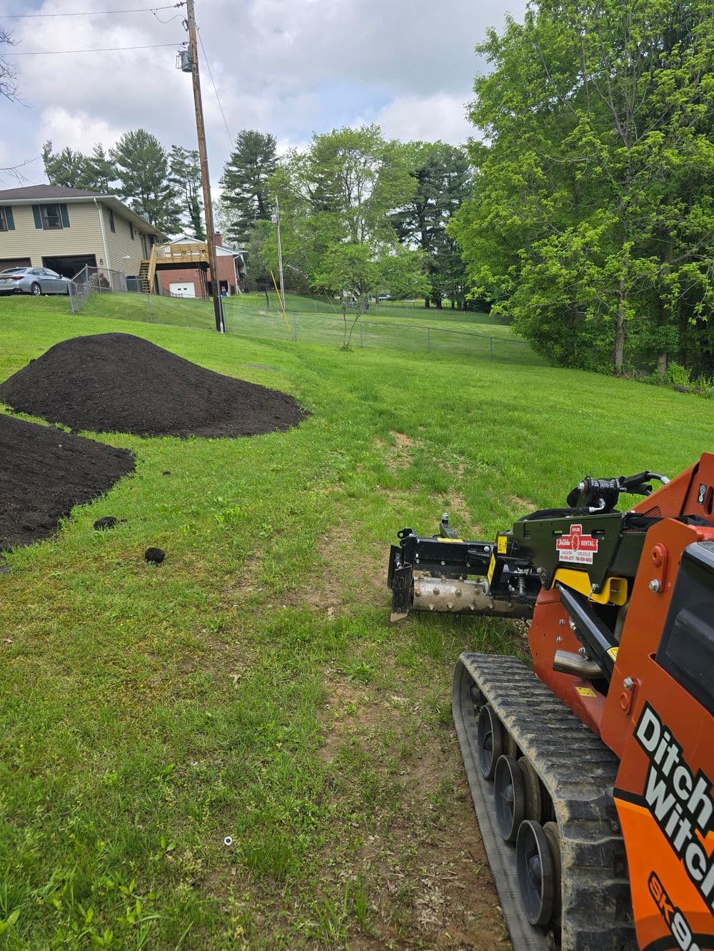 Mini skid steer loader on a grassy lawn near a dirt pile and residential home.