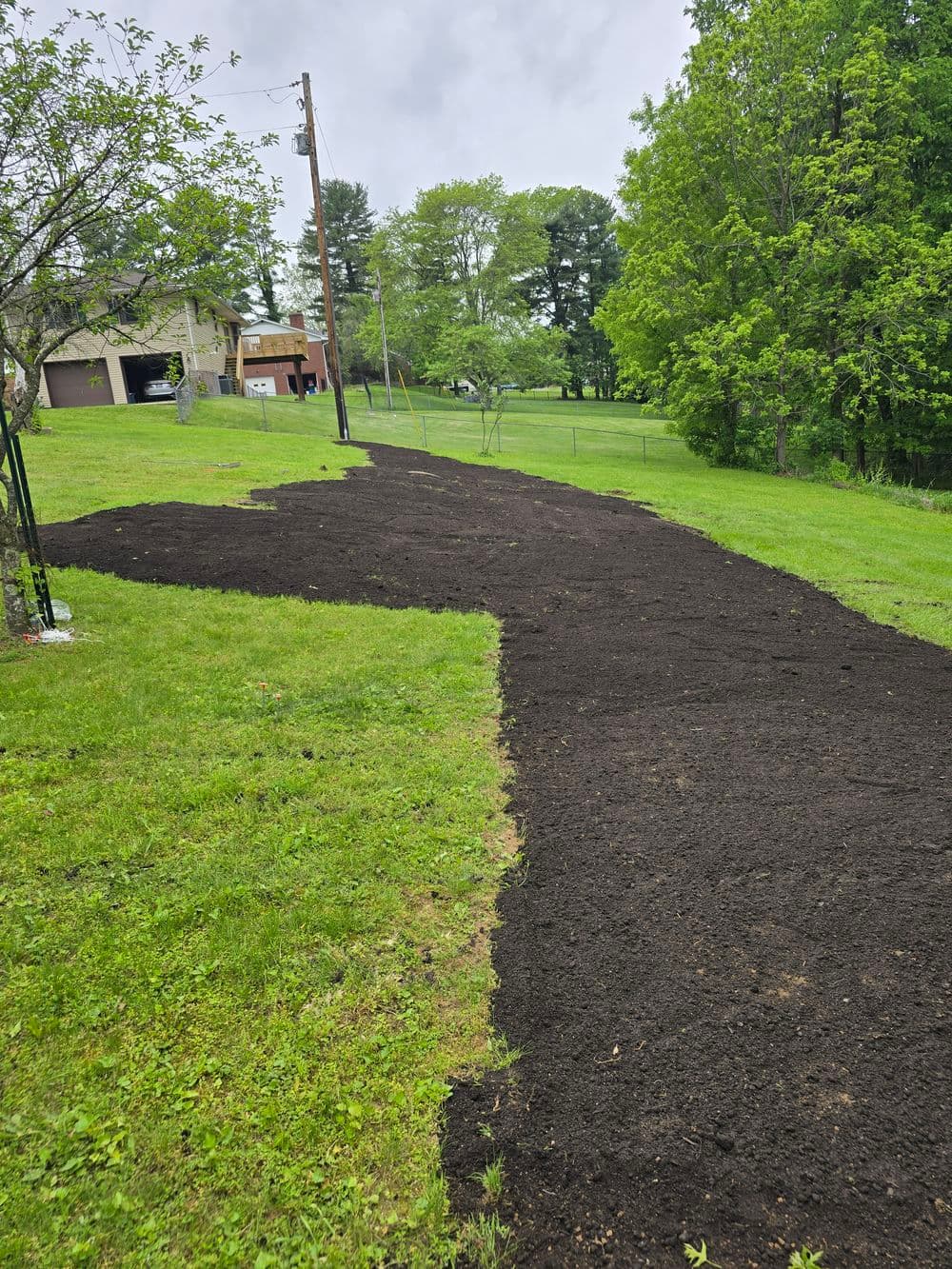 Freshly tilled garden soil path in a green yard, lined with trees and a house in the background.