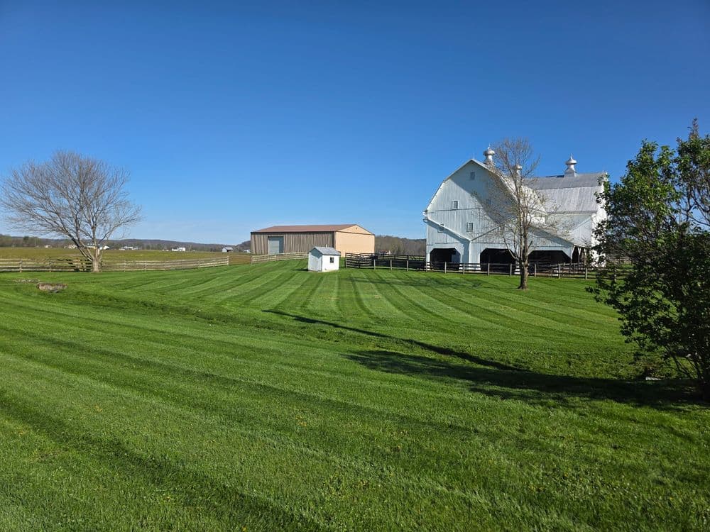 Green lawn with striped grass foreground, farmhouse barn and storage building in the background.