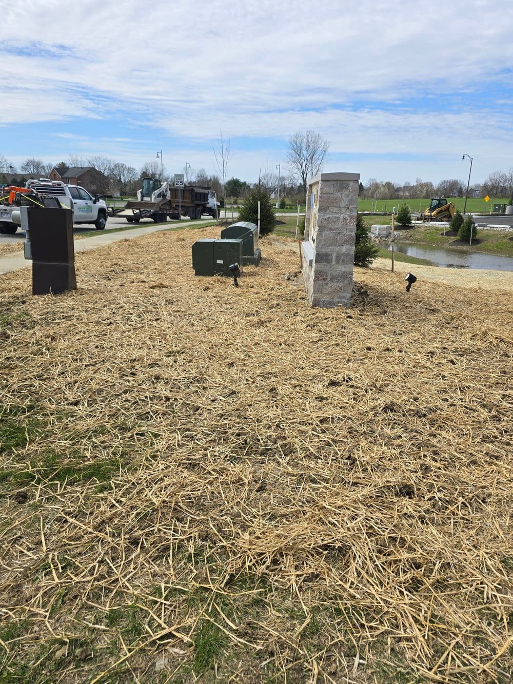 Landscape with straw mulch covering soil, utility boxes, and a stone sign under a blue sky.